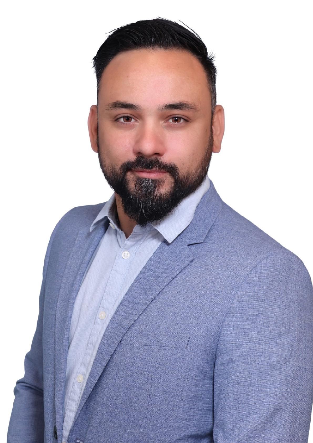 Man with a beard wearing a light blue suit jacket and shirt, posing against a white background.