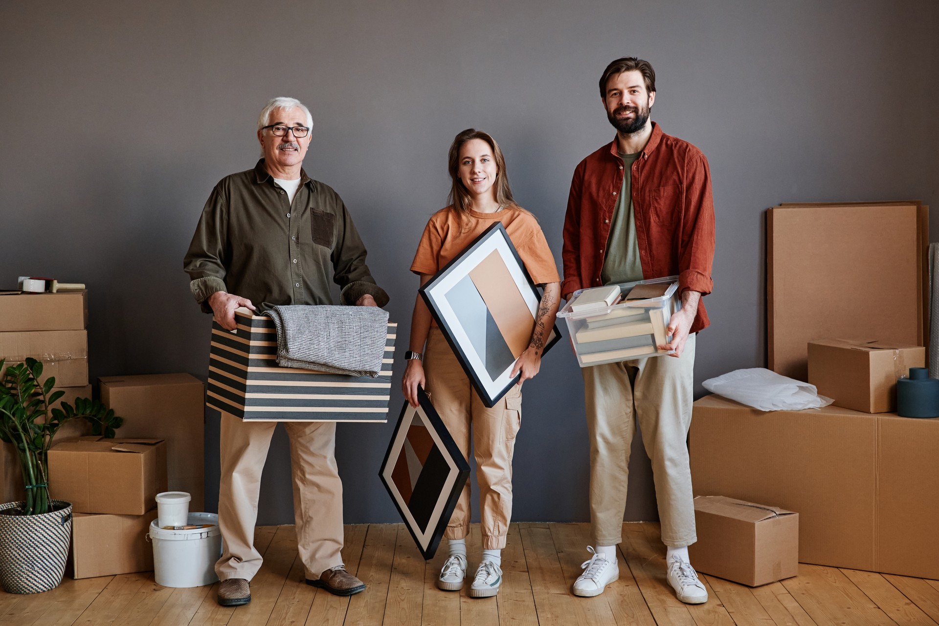 Horizontales Gruppenporträt eines älteren Mannes, seines Sohnes mit seiner Freundin, der zusammen im Loft-Wohnzimmer in einer modernen Wohnung steht und verschiedene Sachen hält